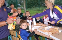 grupa dzieci przy stole oraz kobieta i mezczyzna ktorzr nalewaja sok, pomagaja nalozyc pizze