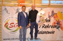 Zawodnik na podium z obok stojącym mężczyzną w eleganckim stroju 
