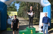 Mężczyzna na podium. Obok dwoje mężczyzn, którzy wręczają nagrody 